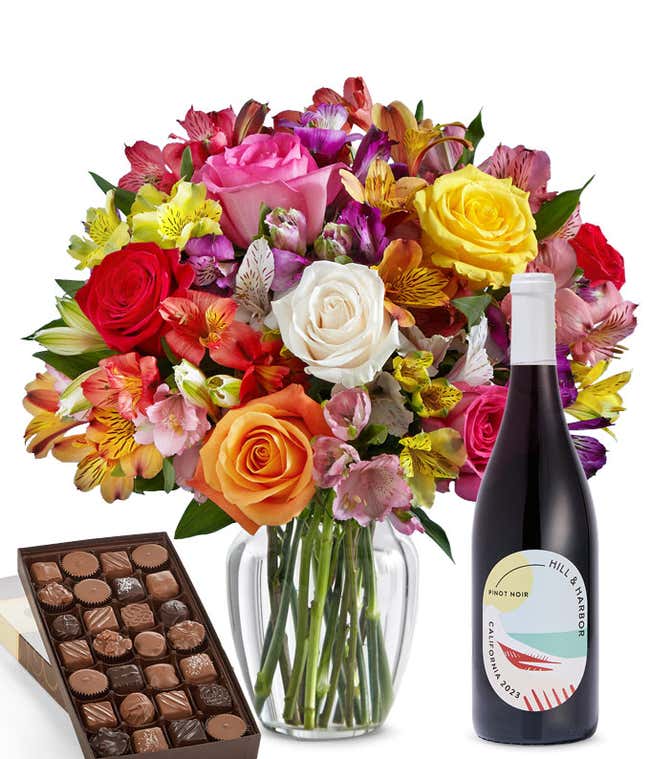 A colorful mixed bouquet of roses and alstroemeria flowers arranged in a clear vase is displayed beside a bottle of Hill &amp; Harbor Pinot Noir and a box of assorted chocolates.