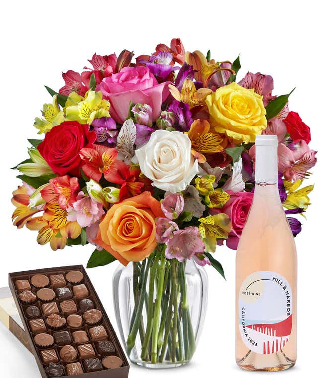 A colorful mixed bouquet of roses and alstroemeria flowers arranged in a clear vase is displayed beside a bottle of Hill &amp; Harbor Ros&eacute; and a box of assorted chocolates.