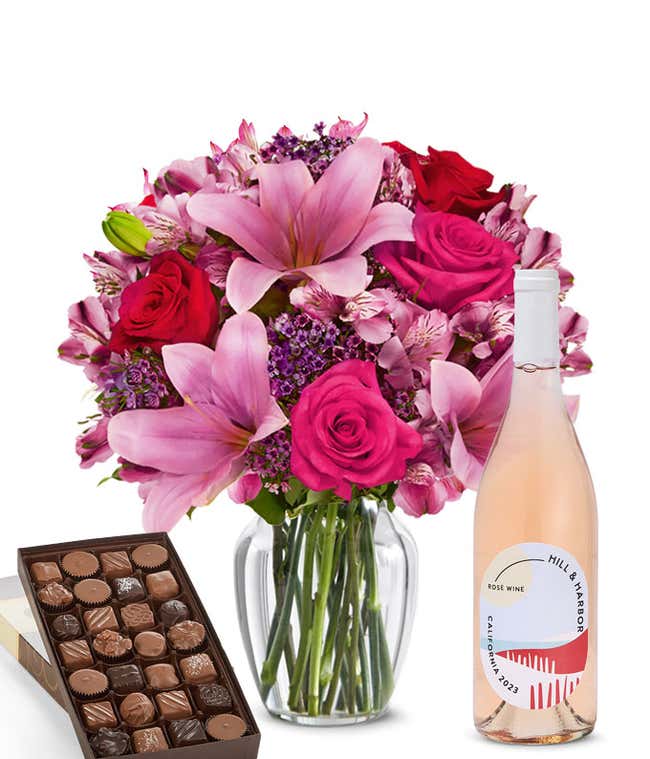 A mixed bouquet of pink and purple flowers in a clear glass vase is paired with a bottle of Hill &amp; Harbor Ros&eacute; and a box of assorted chocolates.