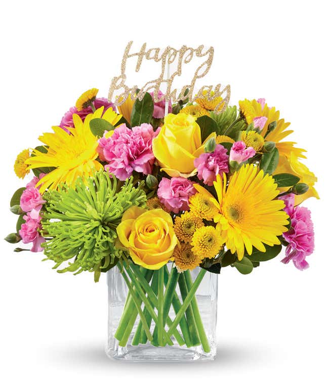 Yellow daisies and roes, pink carns, and green mums with a gold happy birthday pick, in a clear cube vase