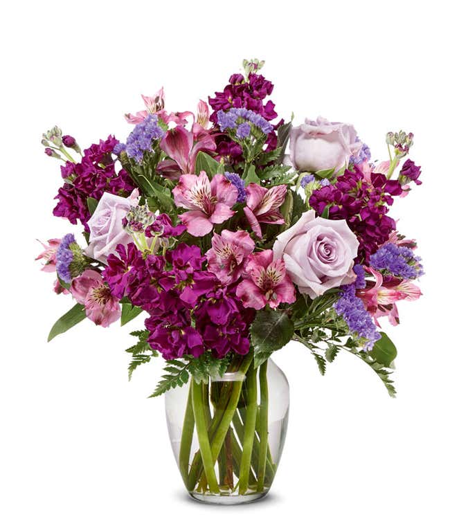 A bouquet of lavender roses, purple alstroemeria and statice, with fresh floral greens, in a clear glass vase 