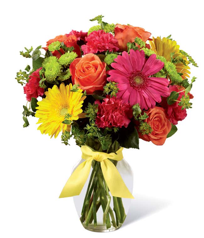 Orange roses, pink and yellow gerbera daisies, with carnations and mixed greens in a clear vase wrapped with a yellow ribbon