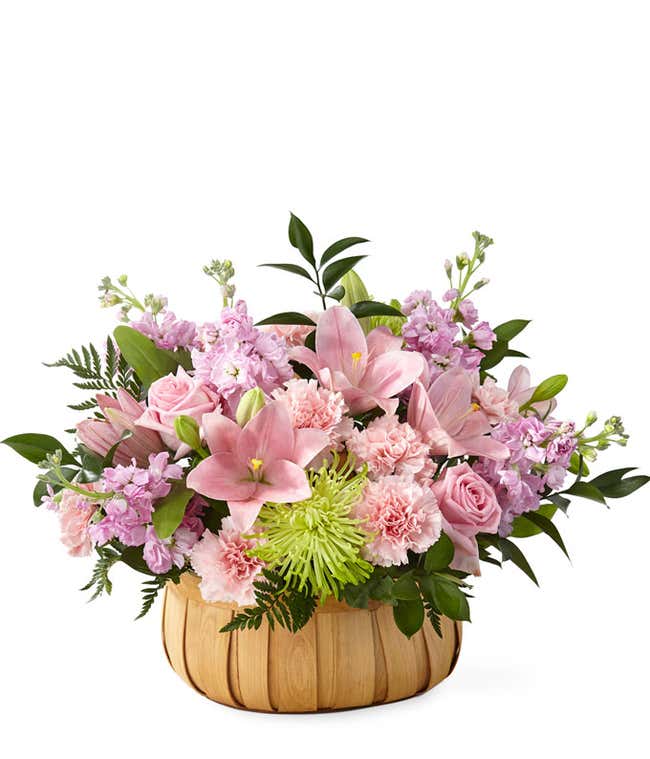 Pink lilies, roes, and carnations, with green poms and fresh floral greens arranged into a wooden basket