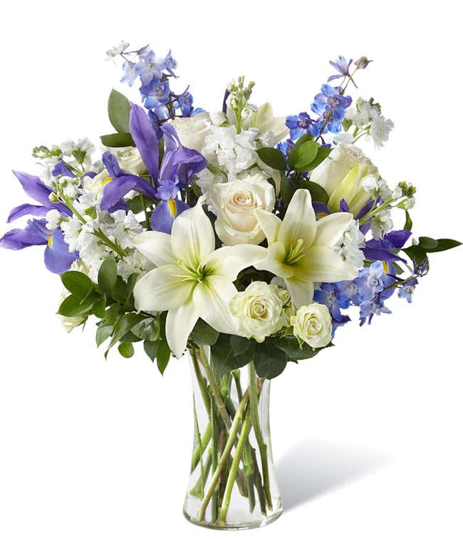 White lilies, blue delphinium, and white roses with fresh floral greens arranged into a tall clear cylinder vase