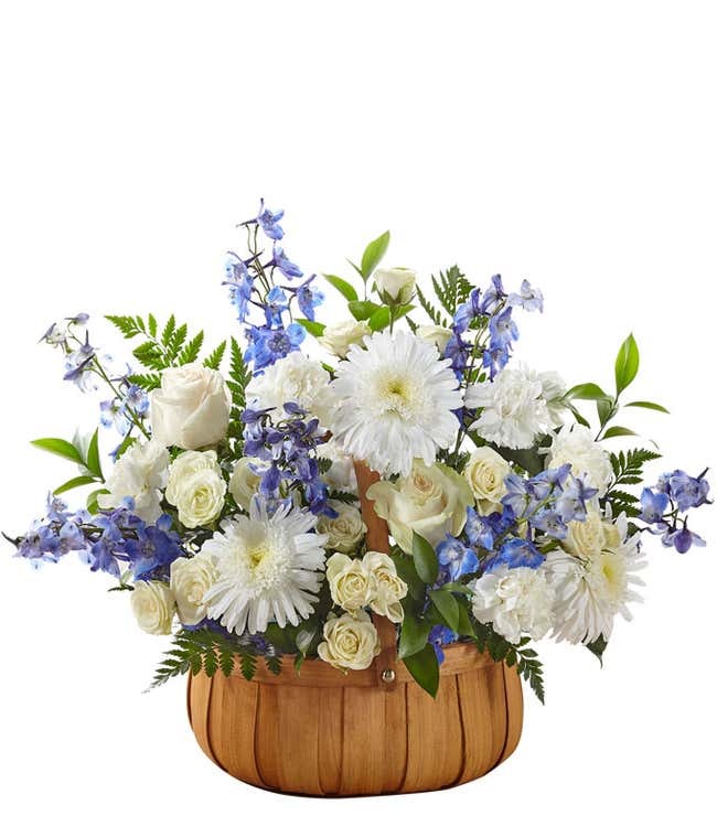 White roses, poms, and carnations, with blue delphinium and fresh floral greens, arranged in a wooden basket