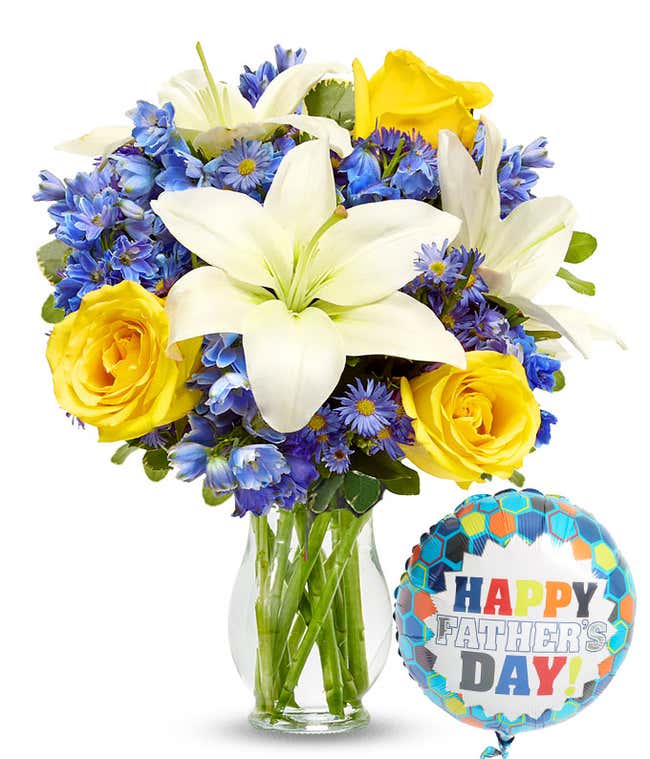White lilies, yellow roses, and tall blue delphinium poke out from a clear vase. Theyr'e accented by variegated green leaves and small purple daisy flowers. Attached is a Happy Father's Day Mylar balloon on a string.