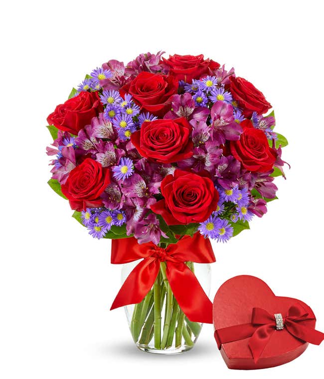 Bouquet of red roses with lots of smaller purple flowers in a clear glass vase with a red bow next to a box of chocoaltes