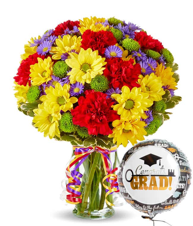 Bouquet of red carnations, yellow daisies, and green and purple filler flowers in a clear vase with colorful curled ribbons and a 'congrats grad!' mylar balloon. 