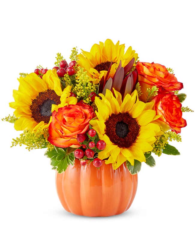  A cheerful floral arrangement featuring sunflowers, orange roses, safari sunset, red berries, and yellow solidago, all beautifully arranged in a pumpkin container.