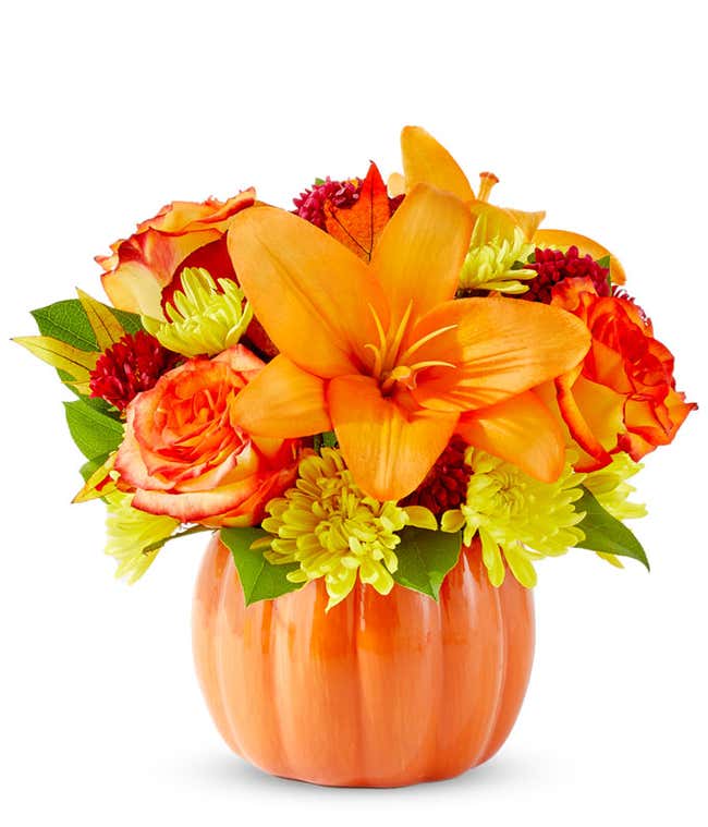  A cheerful floral arrangement featuring orange roses, orange lilies, red Matsumoto asters, and yellow poms, all beautifully arranged in a pumpkin ceramic container.