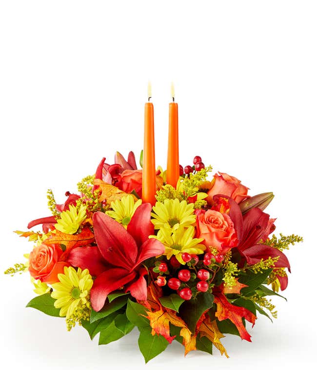 A floral centerpiece of deep red lilies and bright orange roses accented by yellow daisies, red berries, and seeded eucalyptus formed into an oval centerpiece with 2 tall orange tapered candles