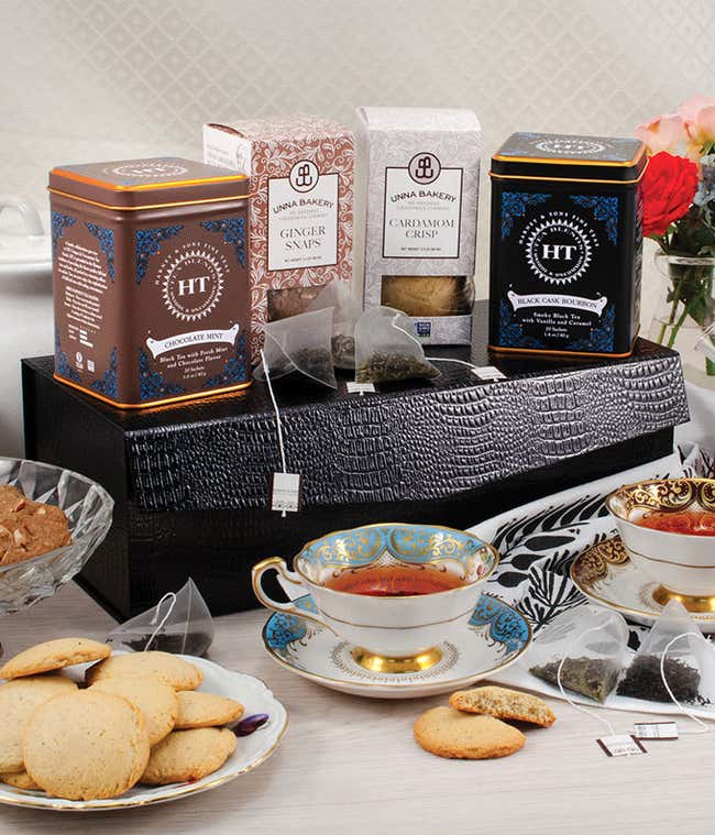 Elegant tea gift set featuring tins of Harney &amp; Sons teas with two ornate teacups filled with tea, surrounded by assorted cookies, packaged tea bags, and a floral arrangement.