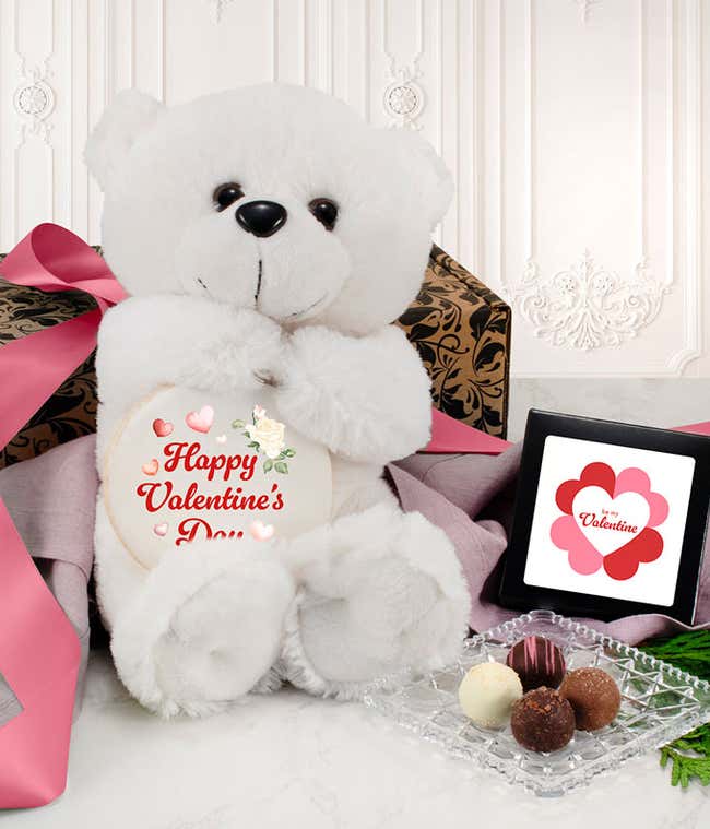 Valentine's Day gift set featuring a white plush teddy bear holding a round sign with a cupid design, a decorative box tied with a pink ribbon, a small framed card with a heart-shaped flower design, and a glass dish displaying assorted chocolate truffles.