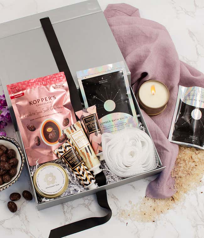 Gift box with bath and spa items including bath salts, body lotion, candle, loofah, and chocolate-covered almonds arranged on marble surface with pink cloth and decorative flowers.