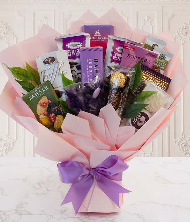 Pink gift bouquet filled with assorted gourmet chocolates, cookies, truffles, and snacks, arranged with greenery and wrapped in blush paper with a purple ribbon bow.