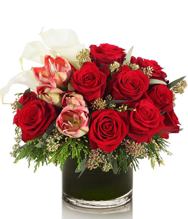  Arrangement of red roses, white calla lilies, and red and white striped amaryllis flowers in a clear glass vase with greenery accents.