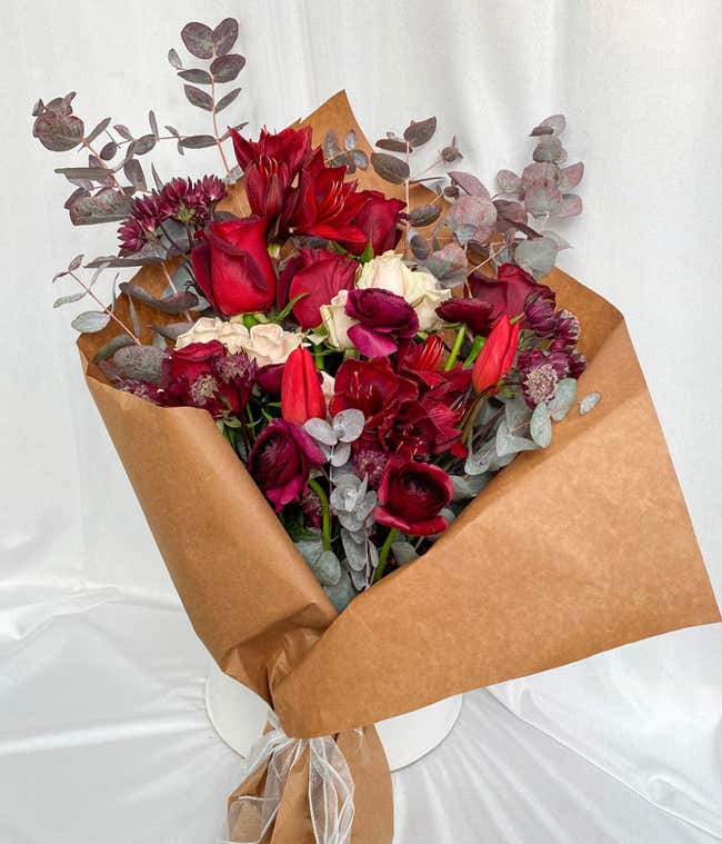 Elegant bouquet wrapped in brown paper featuring a mix of red and white flowers, including roses, tulips, and chrysanthemums, accented with eucalyptus leaves, creating a sophisticated and natural arrangement.