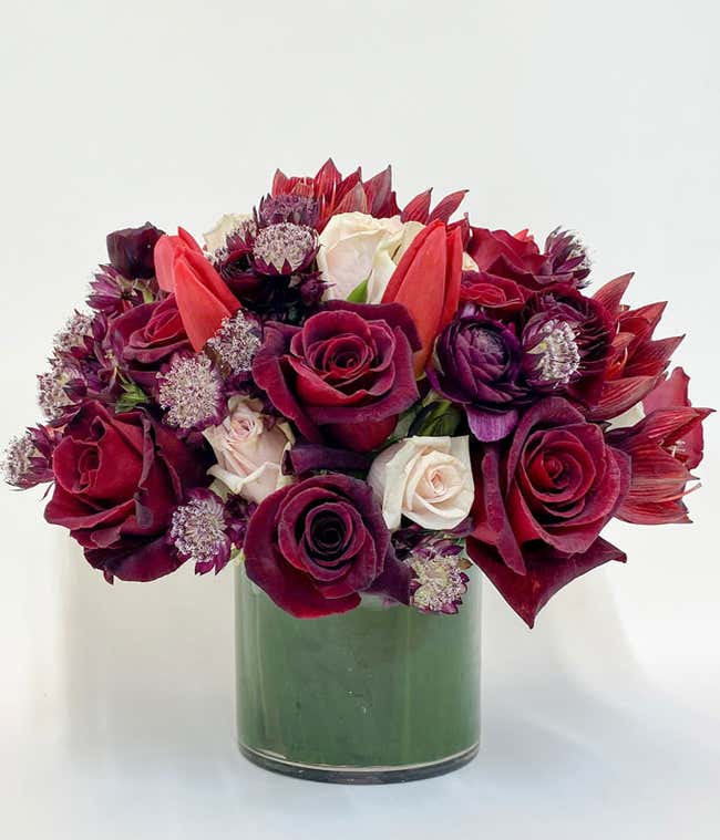 Luxurious bouquet in a clear glass vase featuring a mix of deep red, burgundy, and white flowers, including roses, tulips, and chrysanthemums, arranged with greenery for a rich and elegant floral display.