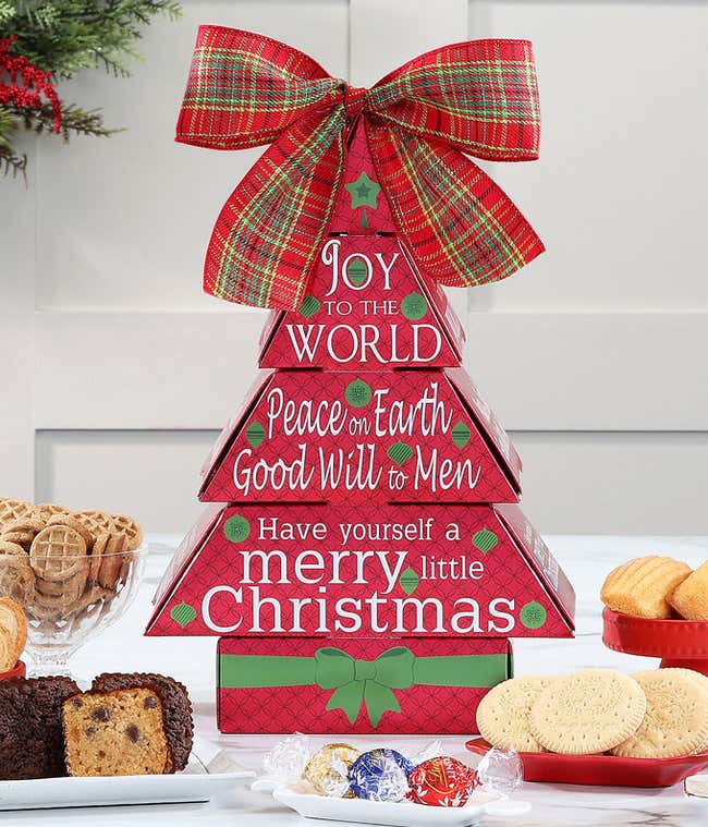Festive gift tower with red and green boxes stacked to form a tree, adorned with holiday greetings like Peace on Earth and Merry Christmas, topped with a white bow with red Ho Ho Ho text. Surrounding treats include Ghirardelli chocolates, cookies, and pep