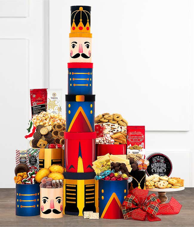 Tall stack of festive nutcracker-themed containers filled with various holiday treats, including cookies, chocolates, popcorn, and candies, displayed with a plaid bow and other seasonal decorations.