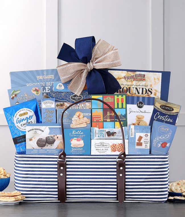  Gift basket with a nautical theme, featuring an assortment of gourmet snacks and treats including chocolates, cookies, crackers, lemon cakes, and mixed olives, all arranged in a blue and white striped tote with leather handles and a blue and white bow.