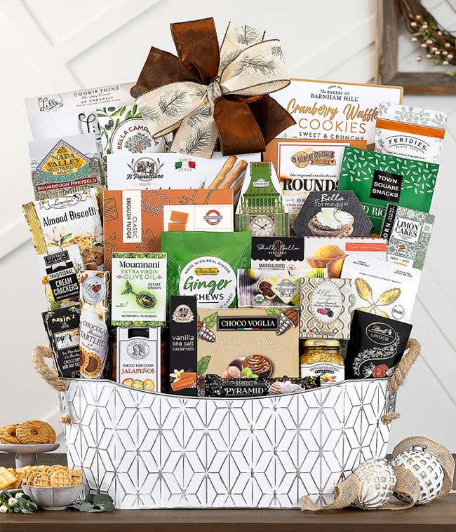 Elegant white patterned gift basket filled with gourmet treats including lemon cake, cheese spread, crackers, pretzels, chocolates, caramels, cookies, tea, and more, topped with a large decorative bow.