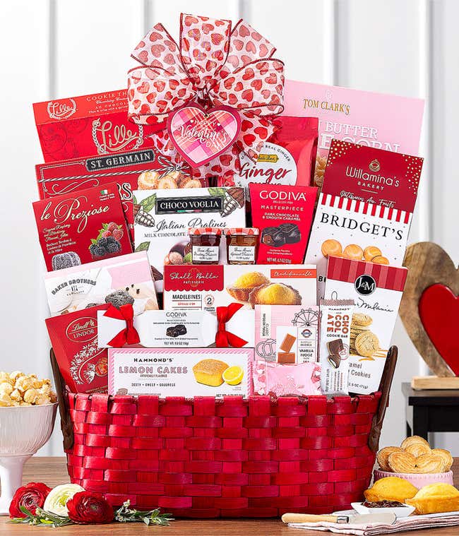 White gift basket filled with assorted gourmet treats in red and pink packaging, including chocolates, cookies, pastries, and popcorn, adorned with a red and pink heart-patterned ribbon.