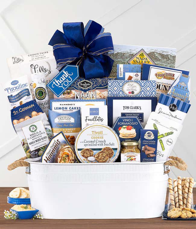 White gift basket with navy blue bow, filled with Ahmad English Tea, cookies, chocolates, hummus, cheese spread, pretzels, olives, and gourmet snacks, displayed with a small blue dish of treats.