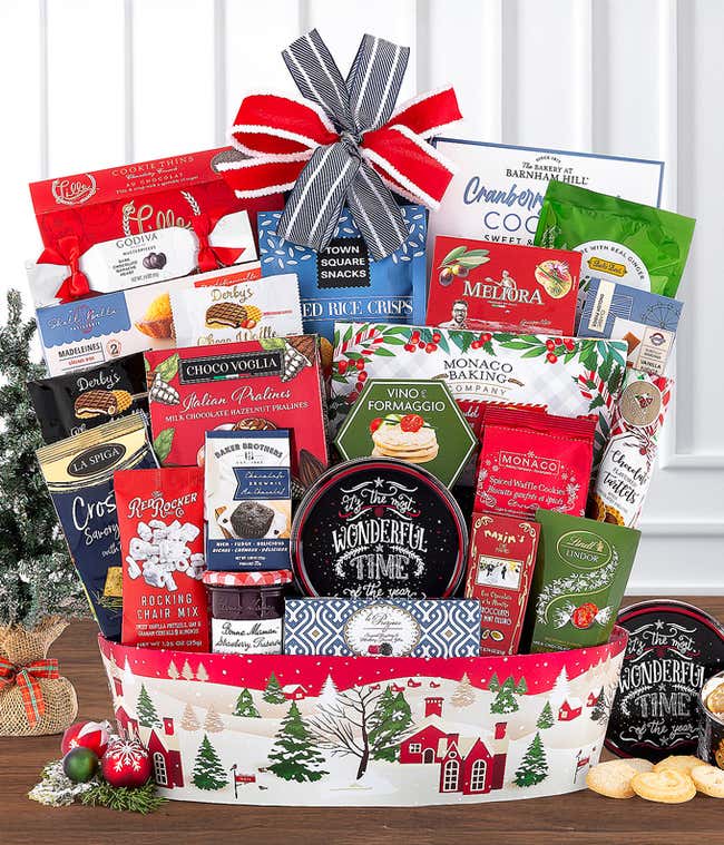 Festive holiday gift basket filled with an assortment of gourmet snacks, cookies, chocolates, and more. Arranged in a red and white Christmas-themed container with village illustrations and topped with a red, white, and navy bow.