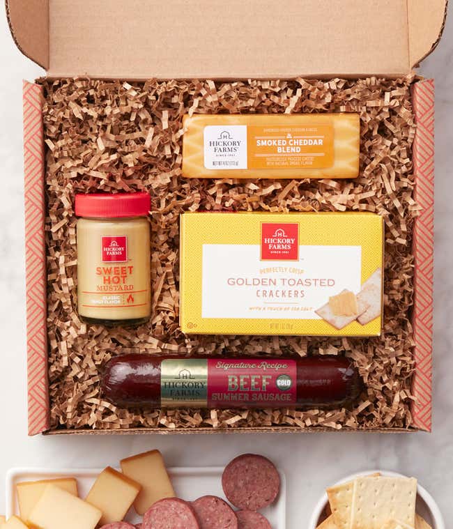 Gift box with assorted savory snacks including beef summer sausage, golden toasted crackers, smoked cheddar blend, sweet hot mustard, and smooth &amp; creamy blend, displayed with sliced sausage, cheese, and crackers on a marble surface.