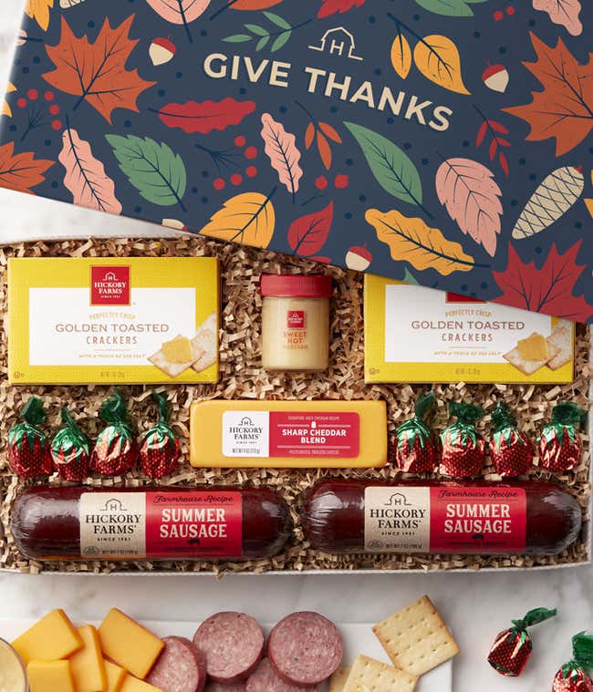Gift box with a fall Give Thanks design, golden toasted crackers, three summer sausages, two cheese blocks, a small jar of sweet hot mustard, and assorted chocolate candies, with a serving tray displaying sliced sausage, cheese cubes, and crackers.