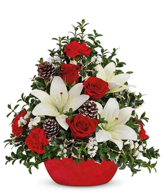 A festive arrangement with red carnations, red roses (deluxe &amp; premium only), white lilies, white sinuata, seasonal greens, a mini Christmas tree, displayed in a red stoneware container