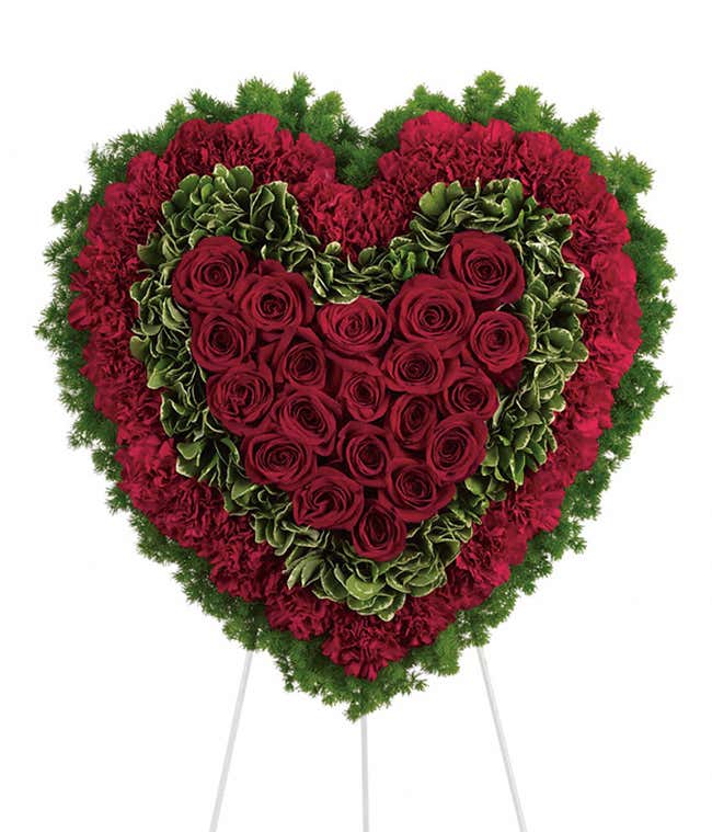 Heart-shaped standing spray with deep red roses, red carnations, and green foliage on a white metal easel.