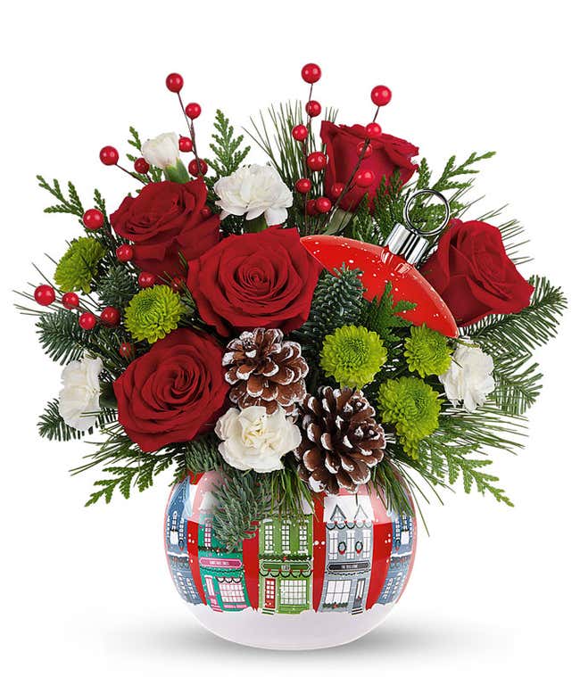 Red roses, white carnations and seasonal greens arranged with berry and pinecone picks in a Christmas village ornament container