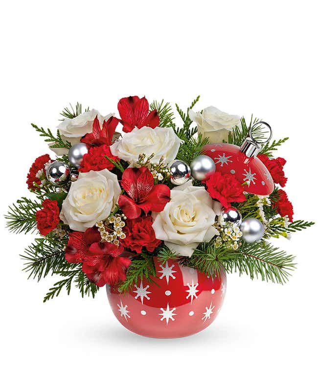 A festive arrangement with red roses, red alstroemeria, red mini carnations, floral greens, silver ornament picks, displayed in a keepsake red ornament vase.