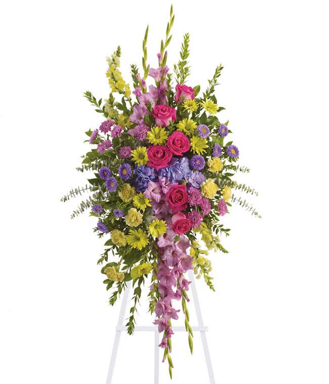 Standing spray with pink roses, purple asters, yellow carnations, lavender gladiolus, and green foliage on a white metal easel.