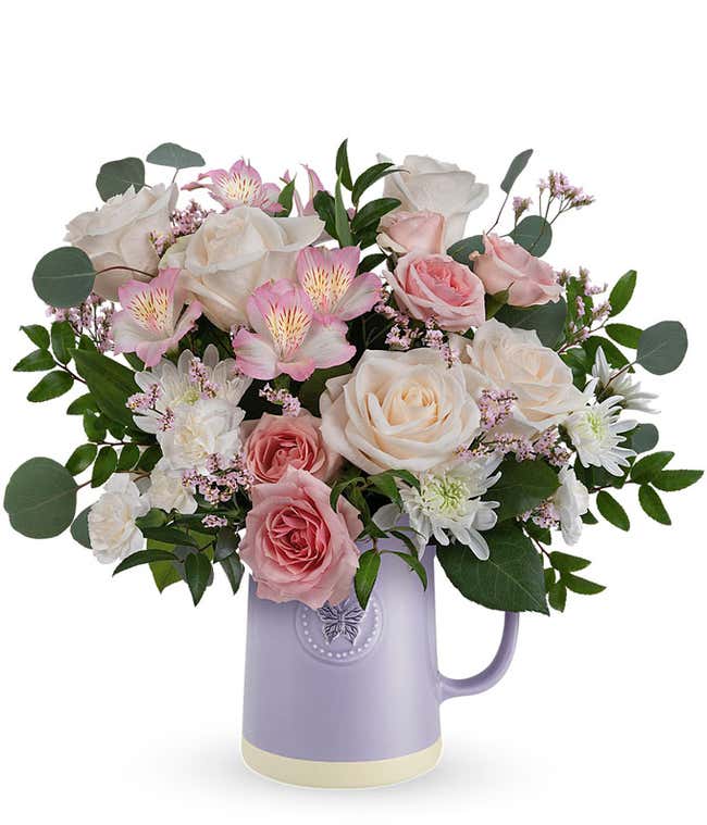 Image of a Mother's Day arrangement featuring Cr&egrave;me Roses, Pink Spray Roses, Light Pink Alstroemeria, White Cushion Spray Chrysanthemums, Pink Limonium, floral greens, all beautifully presented in a keepsake Lavender Butterfly Pitcher.