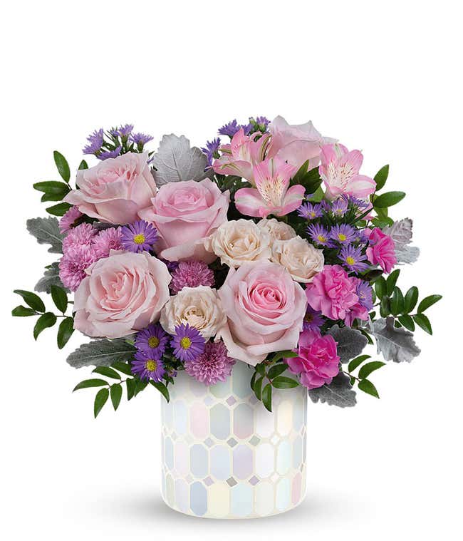 Image of a floral arrangement featuring Pink Roses, Creme Spray Roses, Pink Alstroemeria, Miniature Hot Pink Carnations, Lavender Button Chrysanthemums, and purple Asters, presented in a keepsake iridescent mosaic vase.