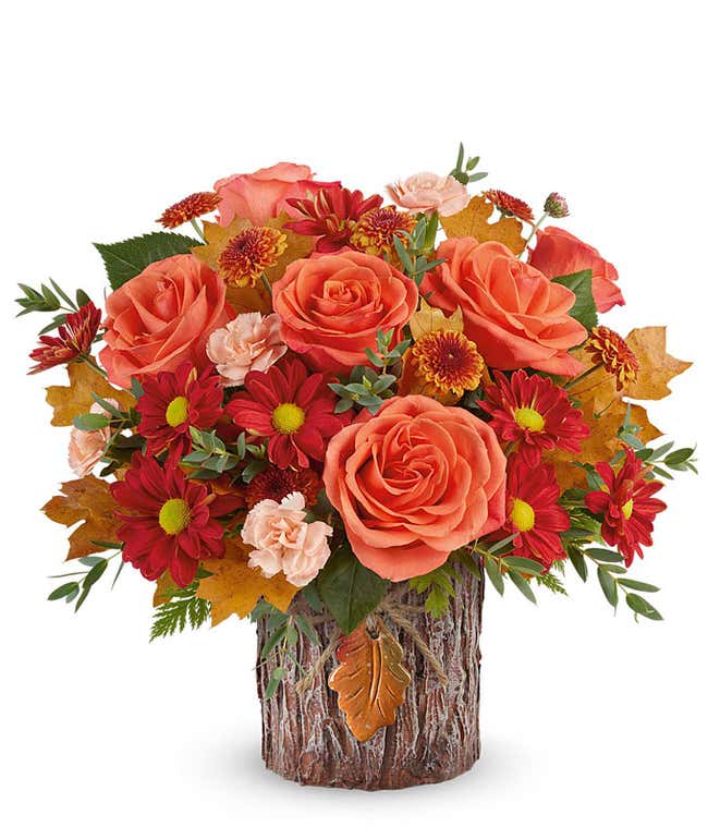 A vibrant arrangement with orange roses, miniature peach carnations, bronze button spray mums, red daisy spray mums, and seasonal greenery in a keepsake autumnal container with a rustic wood texture and leaf accent.