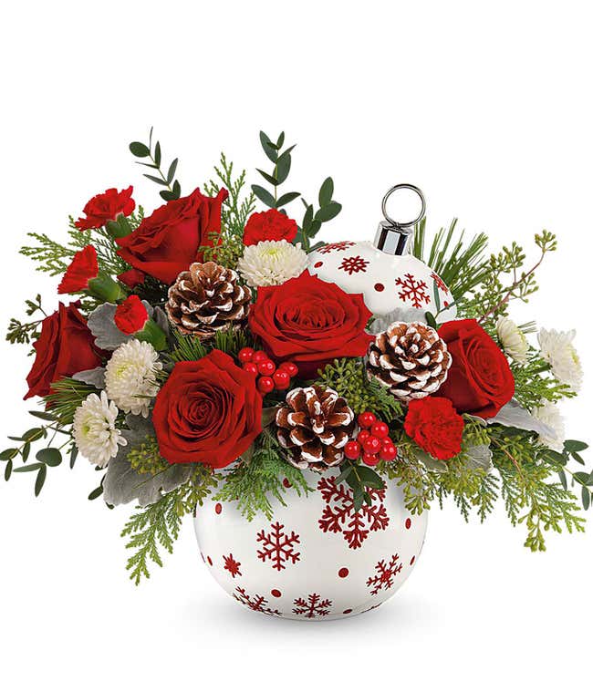 Arrangement with red roses, miniature red carnations, white cushion spray chrysanthemums, pinecones, red berries, and greenery in a white ornament-shaped container decorated with red snowflakes and a silver ornament hook, perfect for the holiday season.