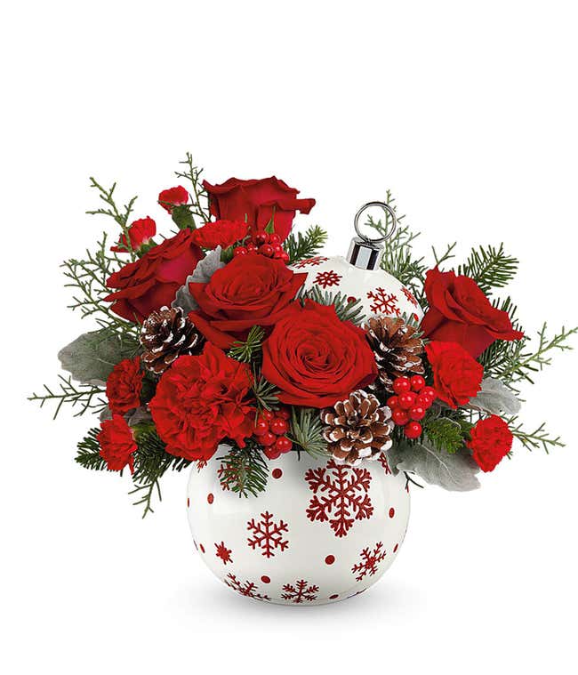 A festive floral arrangement featuring red roses, miniature red carnations, pinecones, red berries, and seasonal greenery. The flowers are displayed in a white ornament-shaped container decorated with red snowflakes, perfect for holiday celebrations.