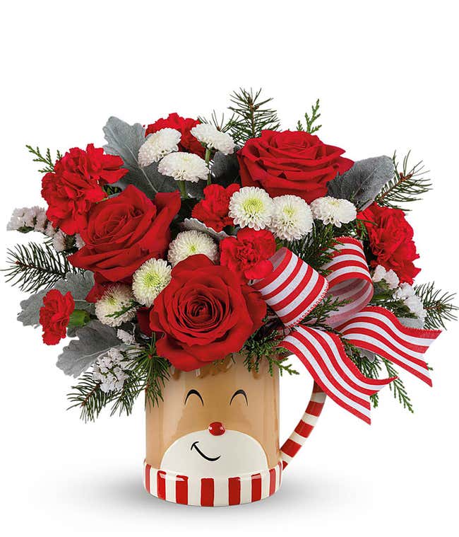 Festive holiday bouquet with red roses, red carnations, white button spray chrysanthemums, dusty miller greenery, and a red and white striped ribbon, arranged in a reindeer-themed mug with a smiling face and striped handle.