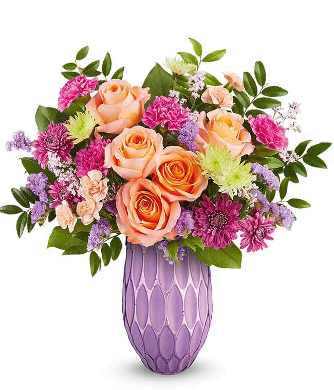 Peach roses, fuchsia carnations, miniature peach carnations, green cushion spray chrysanthemums, purple cushion spray chrysanthemums, purple sinuata statice, and floral greenery arranged in a decorative lavender vase with a textured geometric pattern.