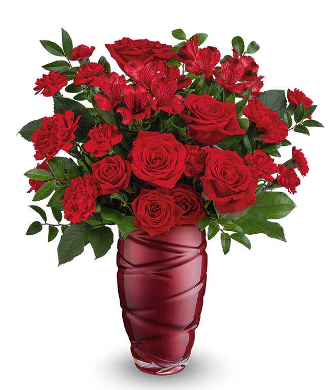 A Valentine's Day bouquet featuring all-red flowers, including roses, carnations, alstroemeria, and lush greenery, arranged in a deep red swirl-patterned glass vase.