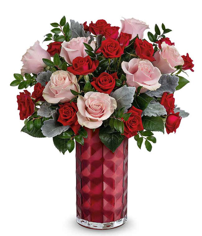 A Valentine's Day bouquet featuring pink roses, red roses, red spray roses, dusty miller, and lush greenery, arranged in a faceted deep red glass vase.