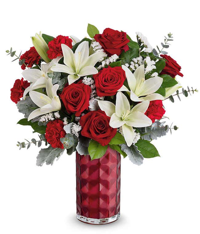 A Valentine's Day bouquet featuring white lilies, red roses, red carnations, white filler flowers, dusty miller, eucalyptus, and lush greenery, arranged in a faceted deep red glass vase.