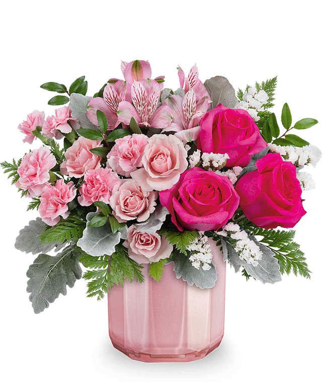 A Valentine's Day bouquet featuring hot pink roses, light pink spray roses, pink alstroemeria, light pink carnations, white filler flowers, dusty miller, and lush greenery, arranged in a soft pink ribbed glass vase.