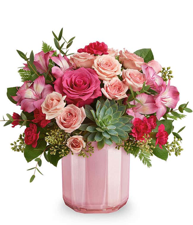 A Valentine's Day bouquet featuring hot pink roses (deluxe and premium sizes only), light pink spray roses, pink alstroemeria, dark pink carnations, a fresh green succulent, seeded eucalyptus, and lush greenery, arranged in a soft pink ribbed glass vase