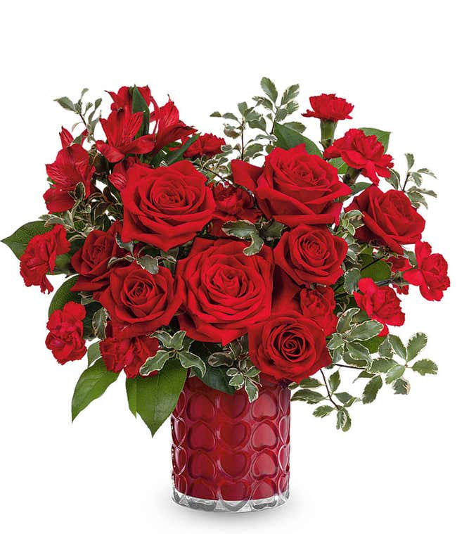 A Valentine's Day bouquet featuring red roses, red spray roses, red carnations, red alstroemeria, variegated greenery, and lush foliage, arranged in a red glass vase with a textured heart pattern.