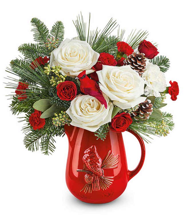 Holiday floral arrangement with white roses, red spray roses, red miniature carnations, pinecones, and greenery in a red ceramic pitcher featuring a raised cardinal design.
